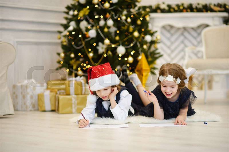Children write letters to Santa Claus. To children it is cheerful. Little boy and girl lie on a floor near Christmas tree. House is festively decorated. Under Christmas tree there are a lot of gifts, stock photo