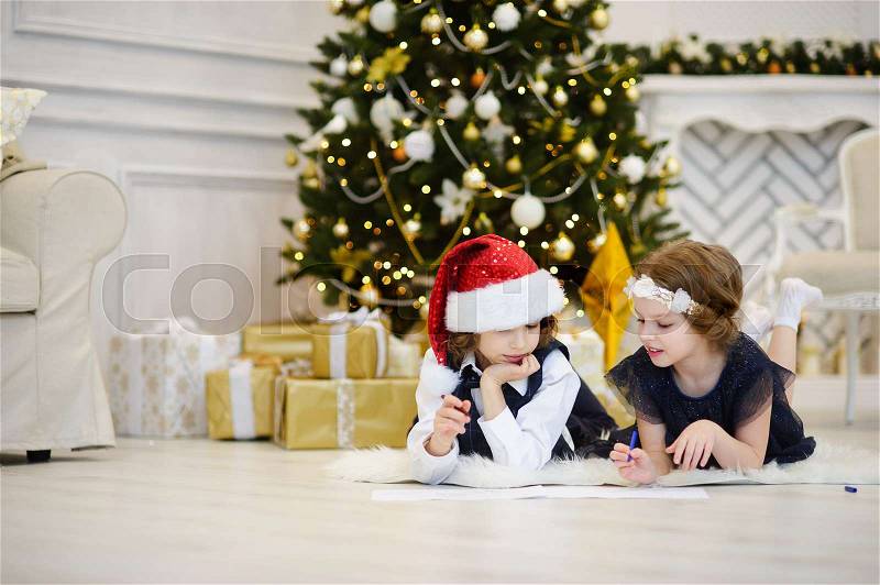 Christmas Eve. Children write letters to Santa Claus. Boy and girl of younger school age lie on a floor near the decorated Christmas tree. Children have thoughtful faces. House is festively decorated, stock photo