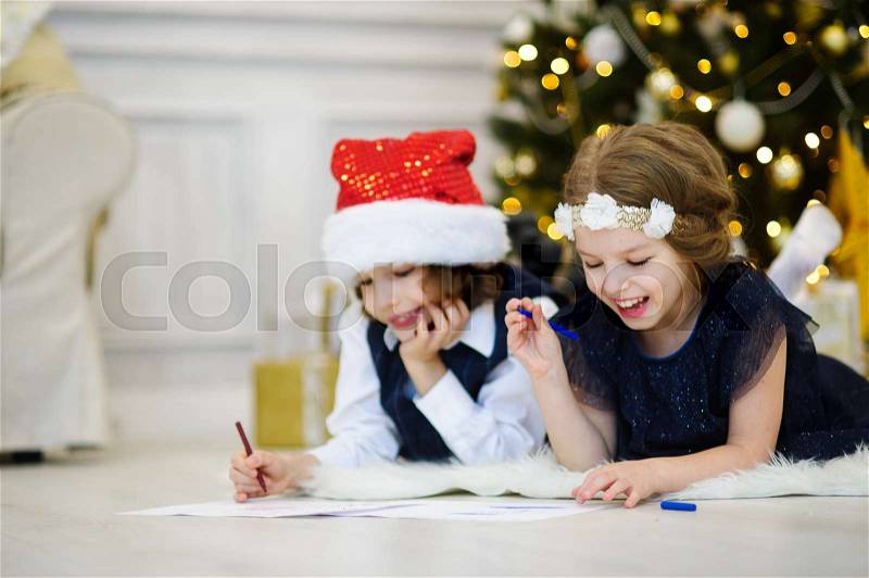 Children write letters to Santa Claus. To children it is cheerful. Little boy and girl lie on a floor near Christmas tree. House is festively decorated. Under Christmas tree there are a lot of gifts, stock photo