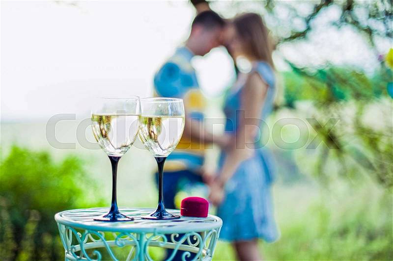 Love, family, anniversary concept - wine glasses and ring box in focus with engaged couple on the background, stock photo