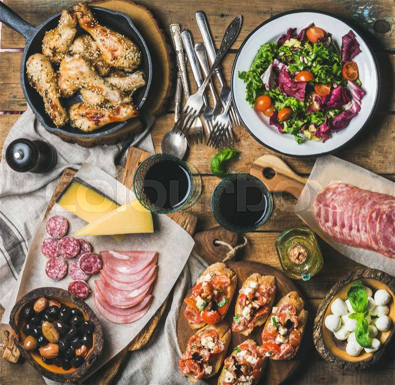 Home dinner, party table arrangement. Rustic table set with salad, olives, chicken, tomato, feta cheese brushettas, snacks and red wine, top view. Slow food, banquet, catering concept, square crop, stock photo