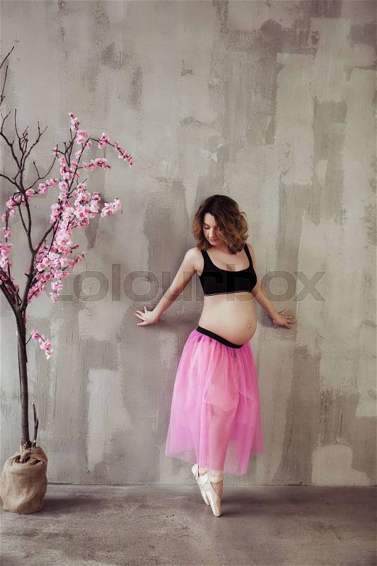 Pregnancy fitness and sport concept happy pregnant woman near the pink sakura tree. Pregnant ballet dancer on gray background. Loft interior, stock photo