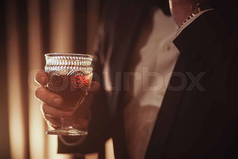 Young ginger bearded man holding a vintage glass with red wine against the light, black on background, stock photo