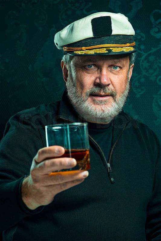 Portrait of old sailor man as captain in black sweater and hat drinking cognac on black studio background, stock photo