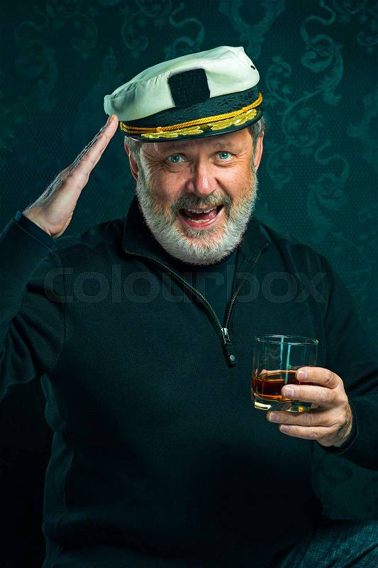 Portrait of old sailor man as captain in black sweater and hat drinking cognac on black studio background, stock photo