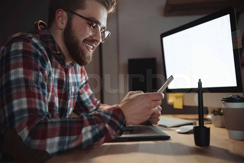 Photo of young happy bearded web designer dressed in shirt in a cage print and wearing glasses chatting by phone late at night, stock photo