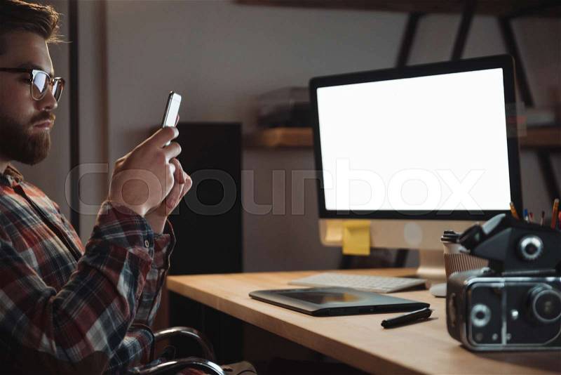 Image of bearded web designer dressed in shirt in a cage print and wearing glasses photographing camera by phone, stock photo