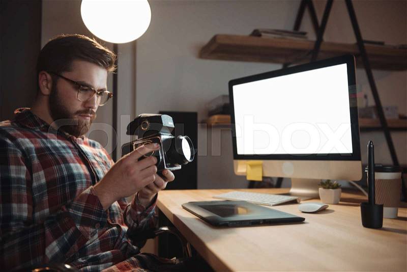 Image of bearded web designer dressed in shirt in a cage print and wearing glasses holding camera. Focus on camera, stock photo