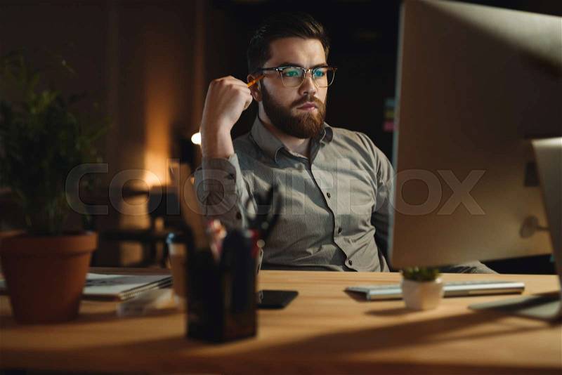 Picture of serious bearded web designer dressed in shirt working late at night and looking at computer while touching head with pencil, stock photo