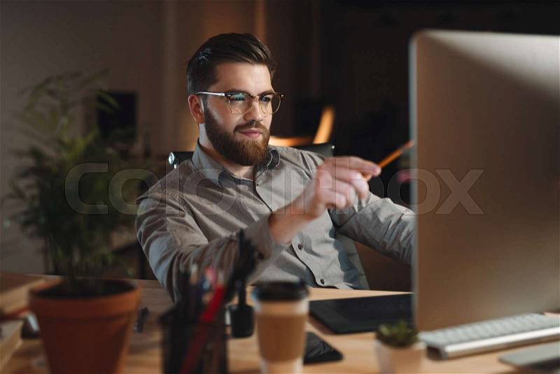 Picture of handsome bearded web designer dressed in shirt working late at night and looking at computer while touching computer display with pencil, stock photo