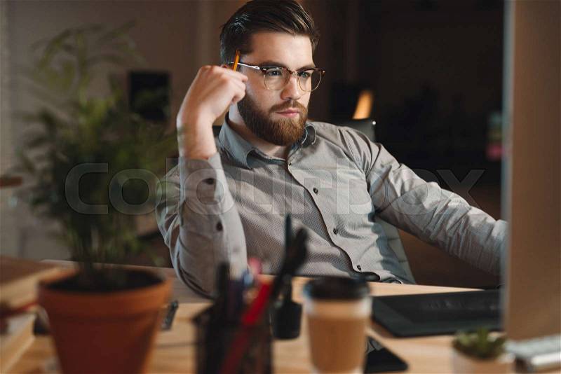 Image of young bearded web designer dressed in shirt working late at night and looking at computer, stock photo