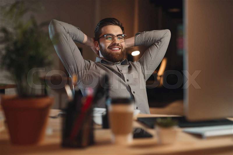 Picture of happy bearded designer dressed in shirt working late at night with computer and stretching, stock photo