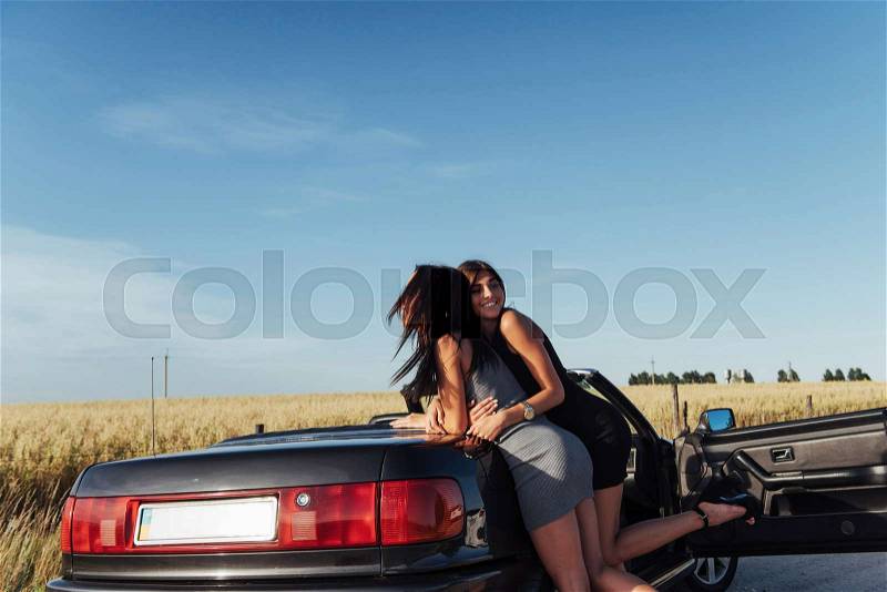 Young women at a photo shoot. Girls gladly posing next to a black convertible against the sky. Summer walk, stock photo