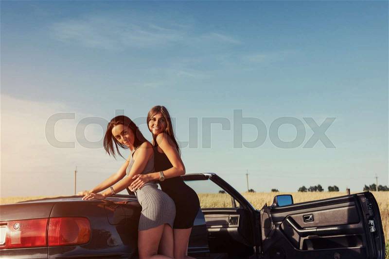 Young women at a photo shoot. Girls gladly posing next to a black convertible against the sky. Summer walk, stock photo