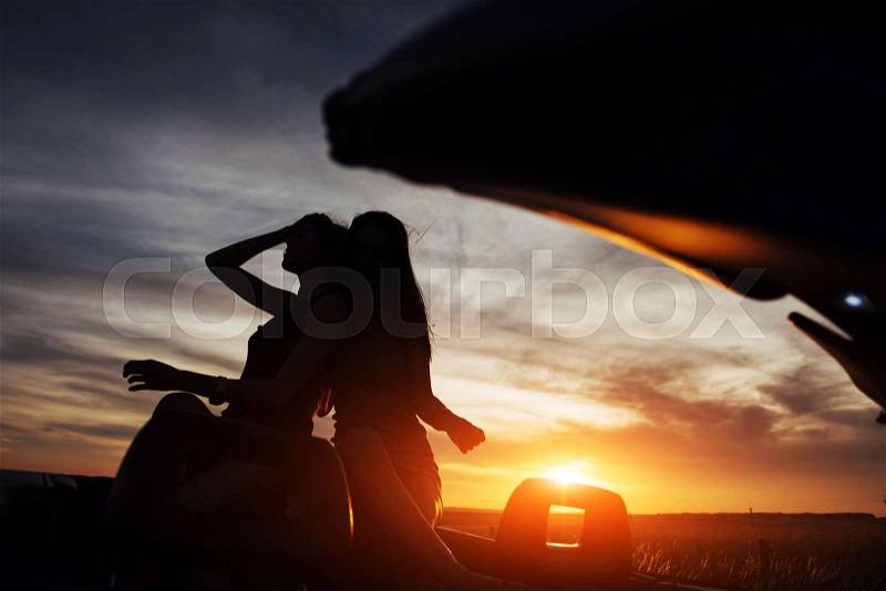 Young two women at a photo shoot. Girls gladly posing next to a black car against the sky on a fantastic sunset, stock photo