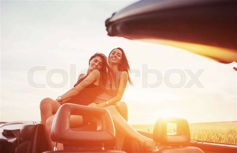 Young two women at a photo shoot. Girls gladly posing next to a black car against the sky on a fantastic sunset, stock photo