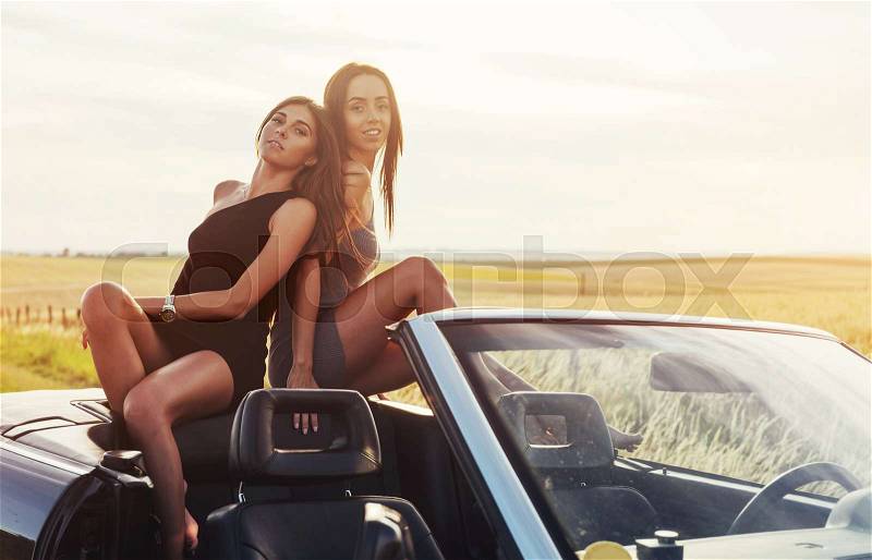 Beautiful two girls are photographed on the road against a background of blue sky and field on a black convertible, stock photo