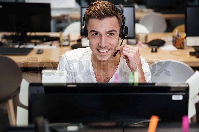 Man sitting by the computer in headphone and looking at camera. Coworking, stock photo
