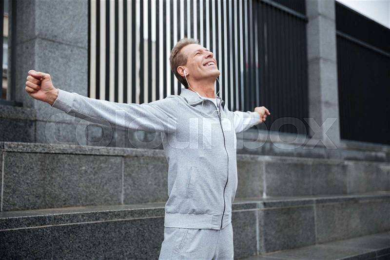 Happy Runner in gray sportswear and headphone warming up on the street with hands to the side. Side view, stock photo