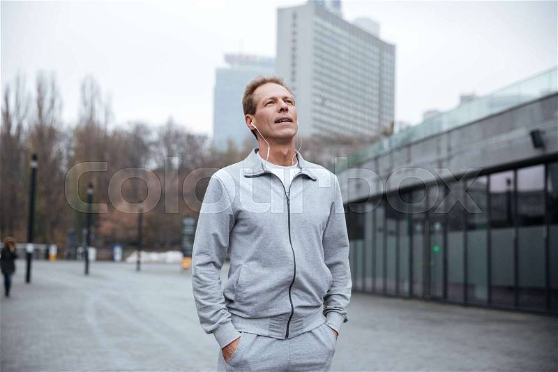 Tired runner in gray sportswear standing on the street with arms in pockets, stock photo