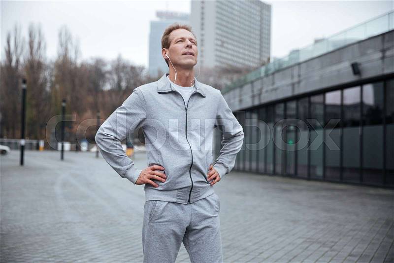 Tired runner in gray sportswear standing on the street with arms at hips, stock photo