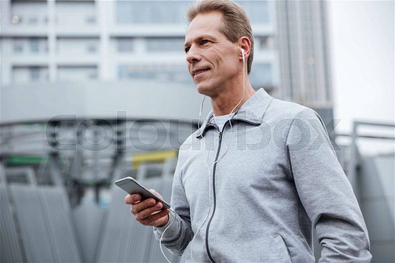 Runner in gray sportswear standing with phone on the street. Side view, stock photo