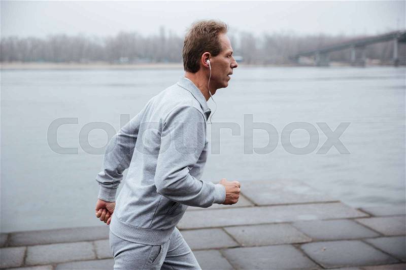 Side view of man in gray sportswear and heaphone running near the water, stock photo