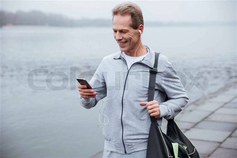 Runner in gray sportswear standing with phone and bag near the river, stock photo