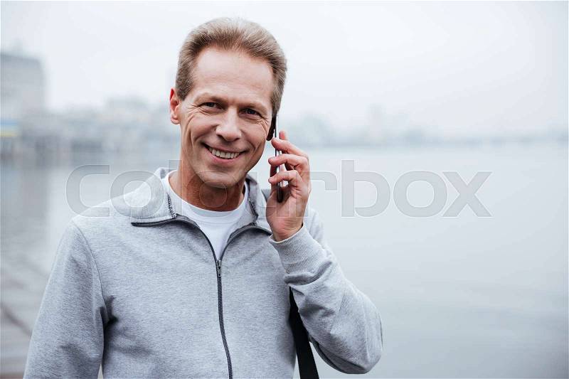 Runner in gray sportswear talking on phone and standing near the river, stock photo