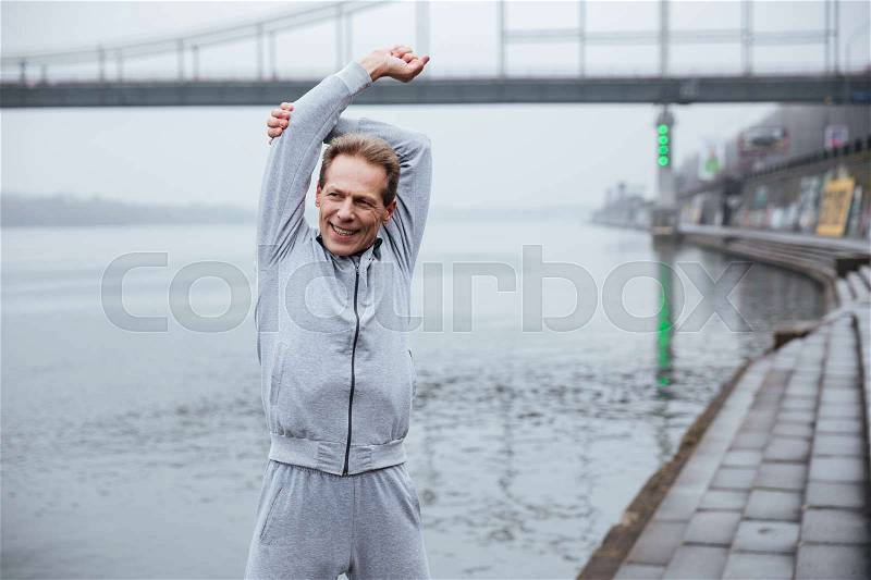 Elderly Smiling man in gray sportswear warming up near the water, stock photo