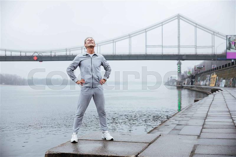 Full length runner in gray sportswear standing near the water with arms at hips, stock photo