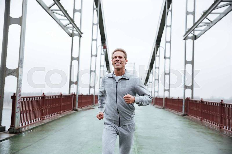 Smiling Man in gray sportswear running on bridge, stock photo