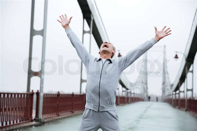 Happy runner in gray sportswear standing on bridge with hands up, stock photo