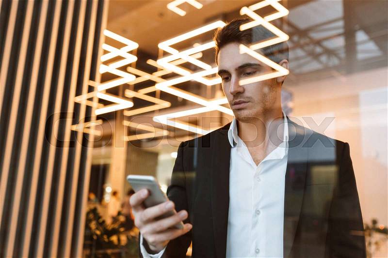 Serious business man in suit behind the glass holding phone in hand and looking at him, stock photo