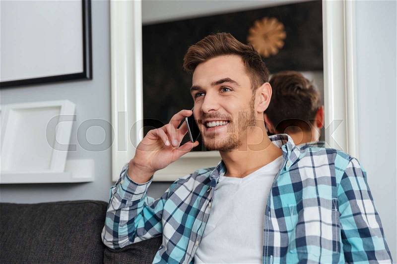 Picture of young happy man dressed in shirt in a cage print sitting on sofa in home and talking by his phone, stock photo