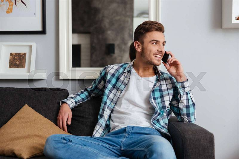 Photo of young cheerful man dressed in shirt in a cage print sitting on sofa in home and talking by his phone. Looking aside, stock photo