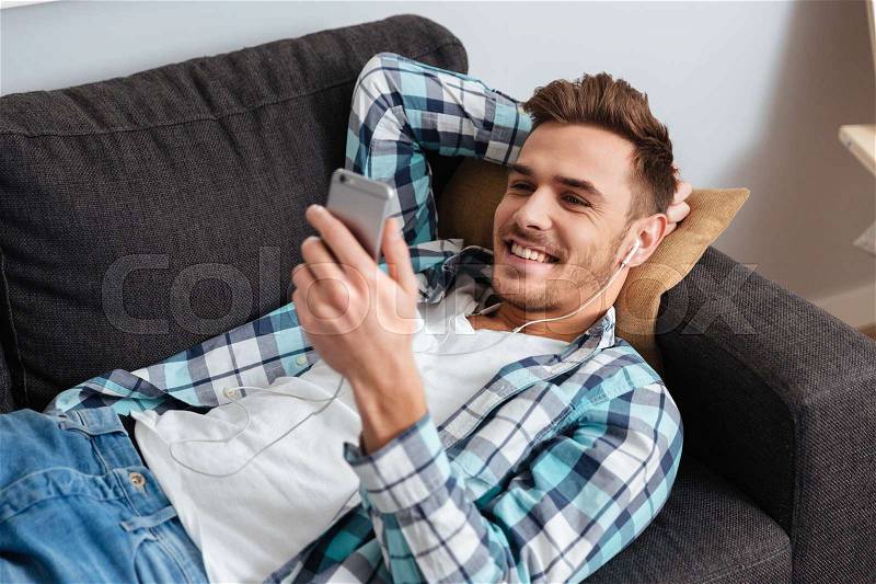 Image of young happy man dressed in shirt in a cage print lies on sofa in home and chatting while listening music with earphones, stock photo