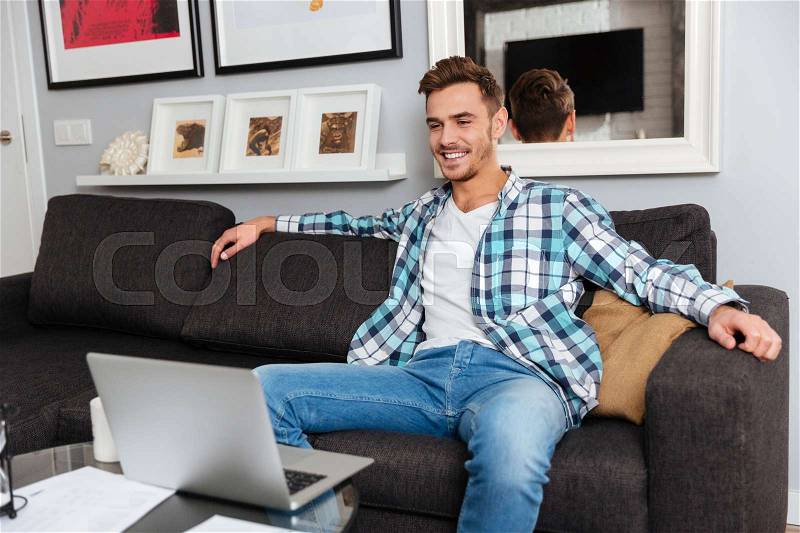Image of happy bristle man dressed in shirt in a cage print sitting on sofa in home and looking at laptop computer, stock photo