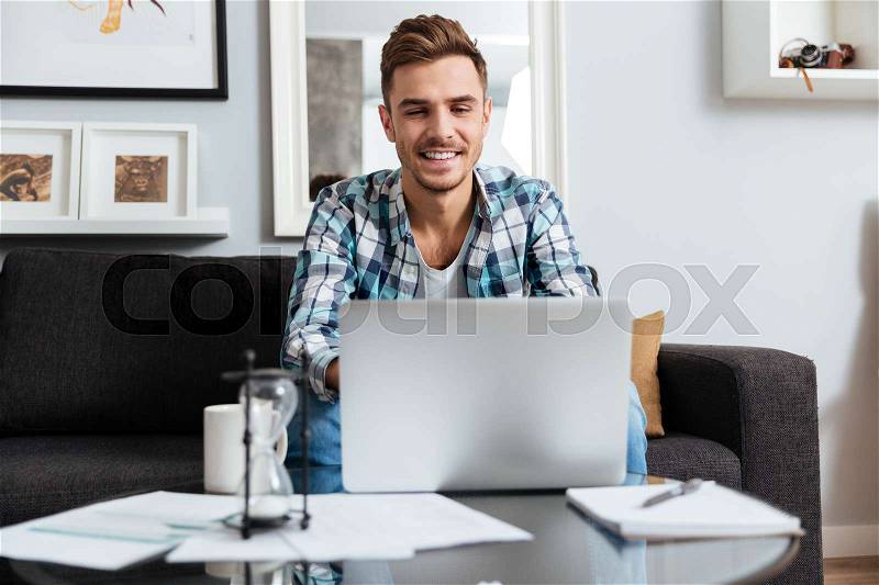 Picture of happy bristle man dressed in shirt in a cage print sitting on sofa in home and looking at laptop computer, stock photo
