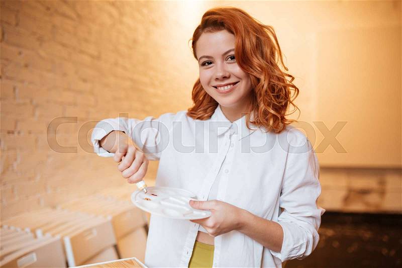 Image of attractive redhead young woman painter with oil paints and palette. Look at camera, stock photo
