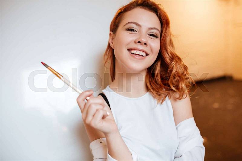 Image of happy attractive young woman painter with red hair standing over blank canvas in artist workshop. Look at camera, stock photo