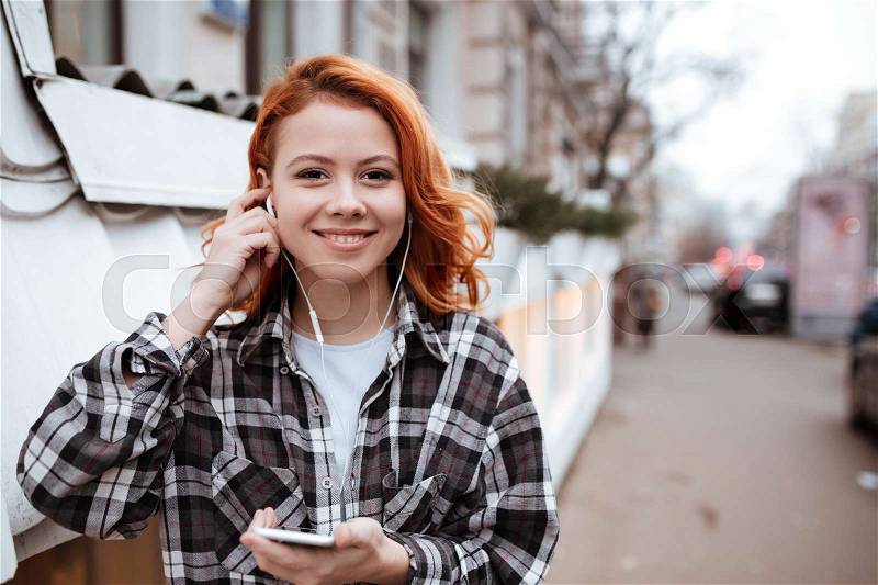 Image of happy young woman with red hair walking outdoors. Look at camera while listen to music and holding phone in hands, stock photo