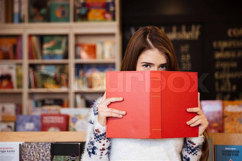Portrait of a young brunette woman covering her face with book with library on background, stock photo