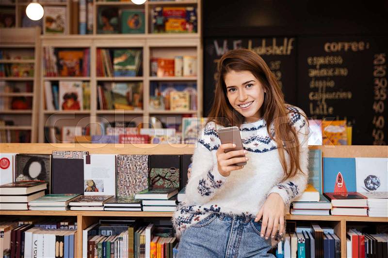 Smiling young woman holding mobile phone while sitting in library, stock photo