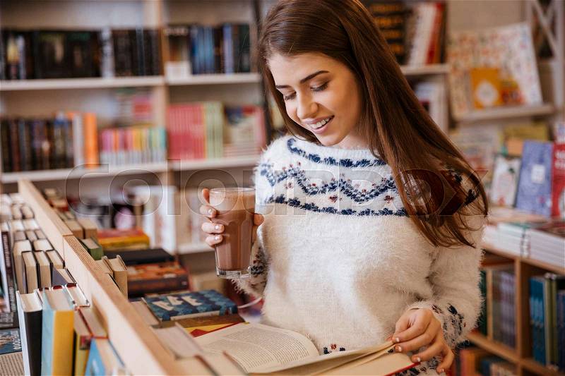 Smiling young woman in sweater reading book and drinking coffee in library, stock photo