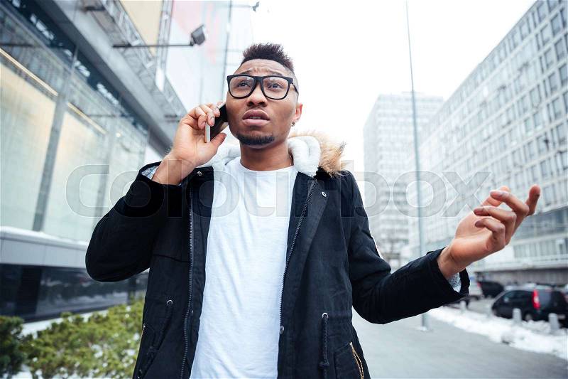 Image of a confused african young man walking on the street while talking by his cellphone. Looking aside, stock photo
