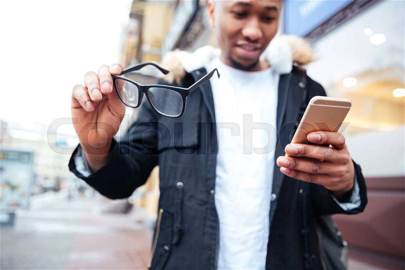Image of african happy man holding his eyeglasses and phone in hands while walking outdoors. Look at phone, stock photo