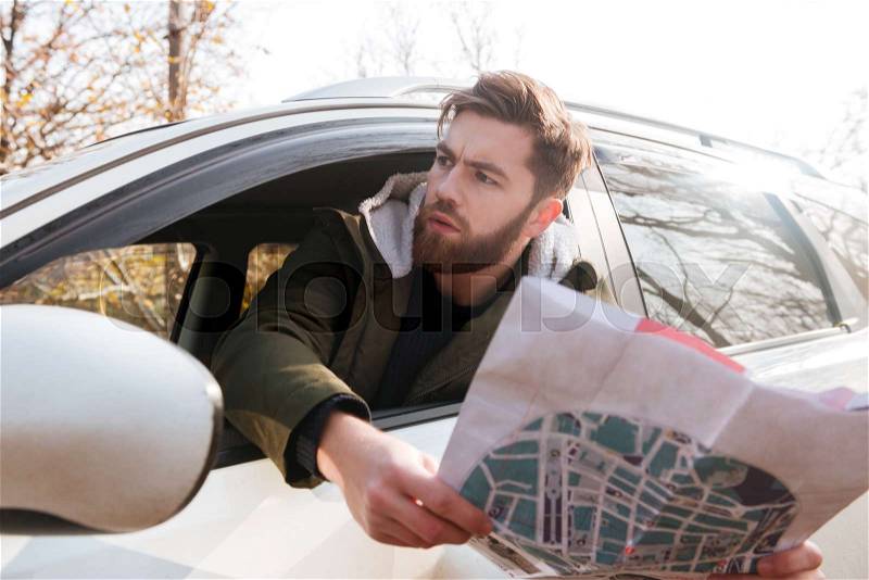 Losted in the forest bearded young driver sitting in car while holding map and looking aside, stock photo