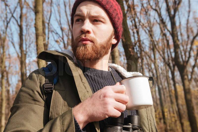 Photo of concentrated bearded man wearing hat holding cup of tea in hands standing in the forest and drinking, stock photo