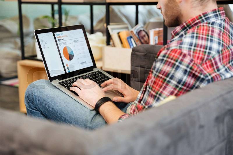 Back view of man analyzes performance on laptop and sitting on sofa in office. Side view, Coworking, stock photo